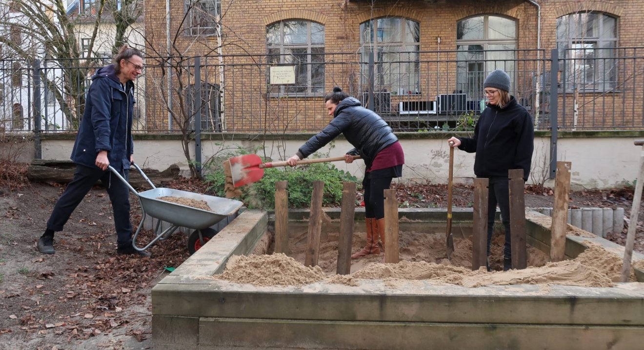 Im Garten des Stadtteilzentrums schippen drei Personen Sand aus dem Sandkasten in eine Schubkarre. 
