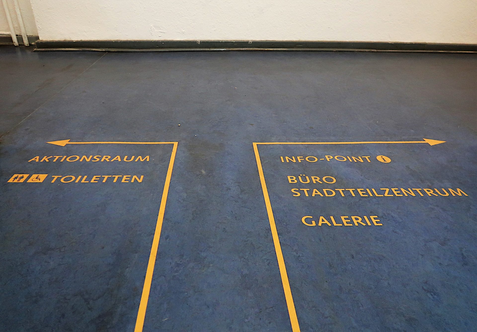 Wege-Leit-System auf dem Fuß-Boden: Ein gelber Pfeil nach Links zeigt Aktions-Raum und Toiletten an, ein Pfeil nach Rechts Info-Point, Büro und Galerie.
