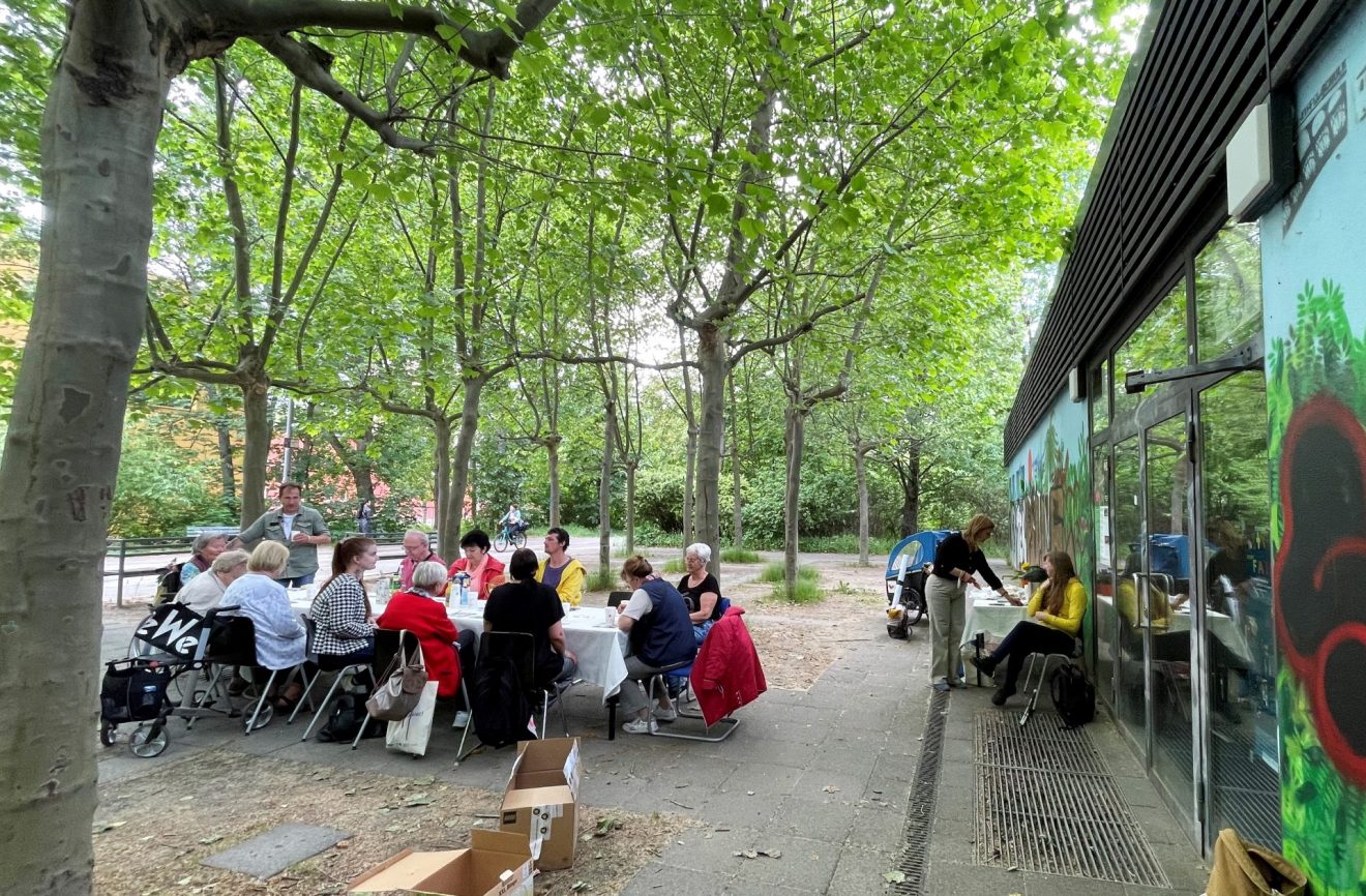 Mehrere Personen sitzen an einem großen Tisch vor dem Quartierspavillon, im Hintergrund viele Bäume.