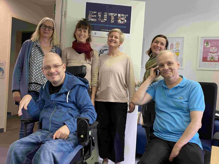 Ein Team-Foto der BZSL. Sechs Personen stehen zusammen im Büro. Vier Frauen stehen im Hintergrund, davor sitzen zwei Männer. Einer von ihnen sitzt in einem elektrischen Rollstuhl, der andere auf einem Schreibtischstuhl.