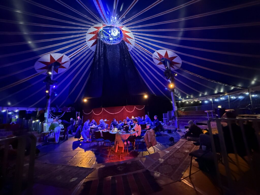 In der MItte des Zirkuszelts sind Esstische aufgebaut. MehrerePersonen sitzen an den Tischen und essen zusammen oder unterhalten sich.