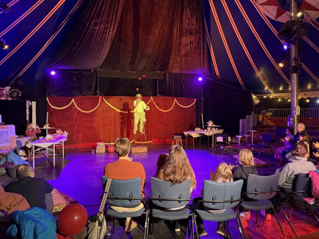 Eine Familie sitzt in der ersten Reihe bei einer Zirkusvorstellung. Vor ihnen jongliert ein Clown auf Stelzen.