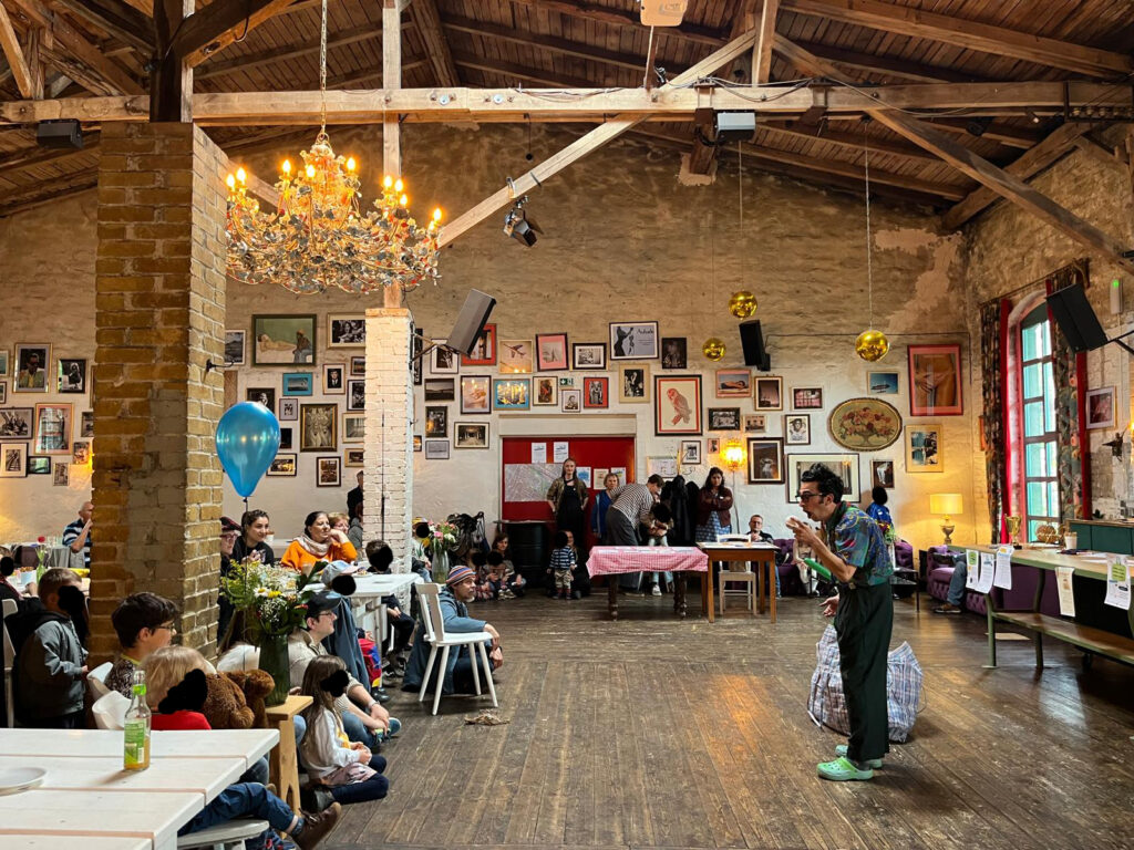 Das Bild zeigt einen Clown während des Kiezfestes. Menschen sitzen auf Stühlen und auf dem Boden. An der Wand hängen verschiedene Bilder.