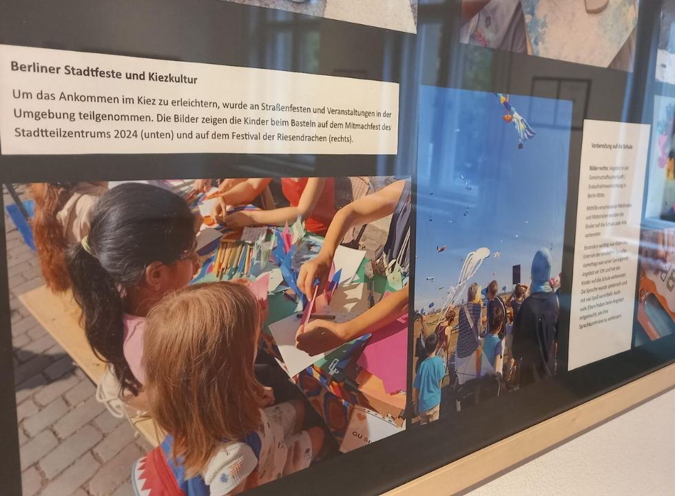 Das Foto zeigt einen Ausschnitt aus der Ausstellung: Ein Foto mit zwei bastelnden Mädchen an einem Markstand. Daneben ein Text zur Erläuterung über das Mitmachfest 2024, bei dem die Kinder aus der Gemeinschaftsunterkunft teilnahmen.