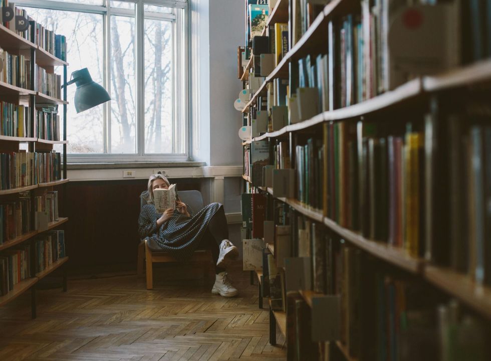 Eine Frau sitzt tief vergraben in einem Sessel in einer Bibliothek und liest in einem Buch