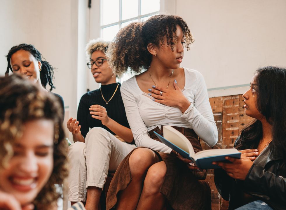 Junge POC-Frauen sitzen in einem Raum und unterhalten sich. Sie halten gemeinsam ein Buch in der Hand.